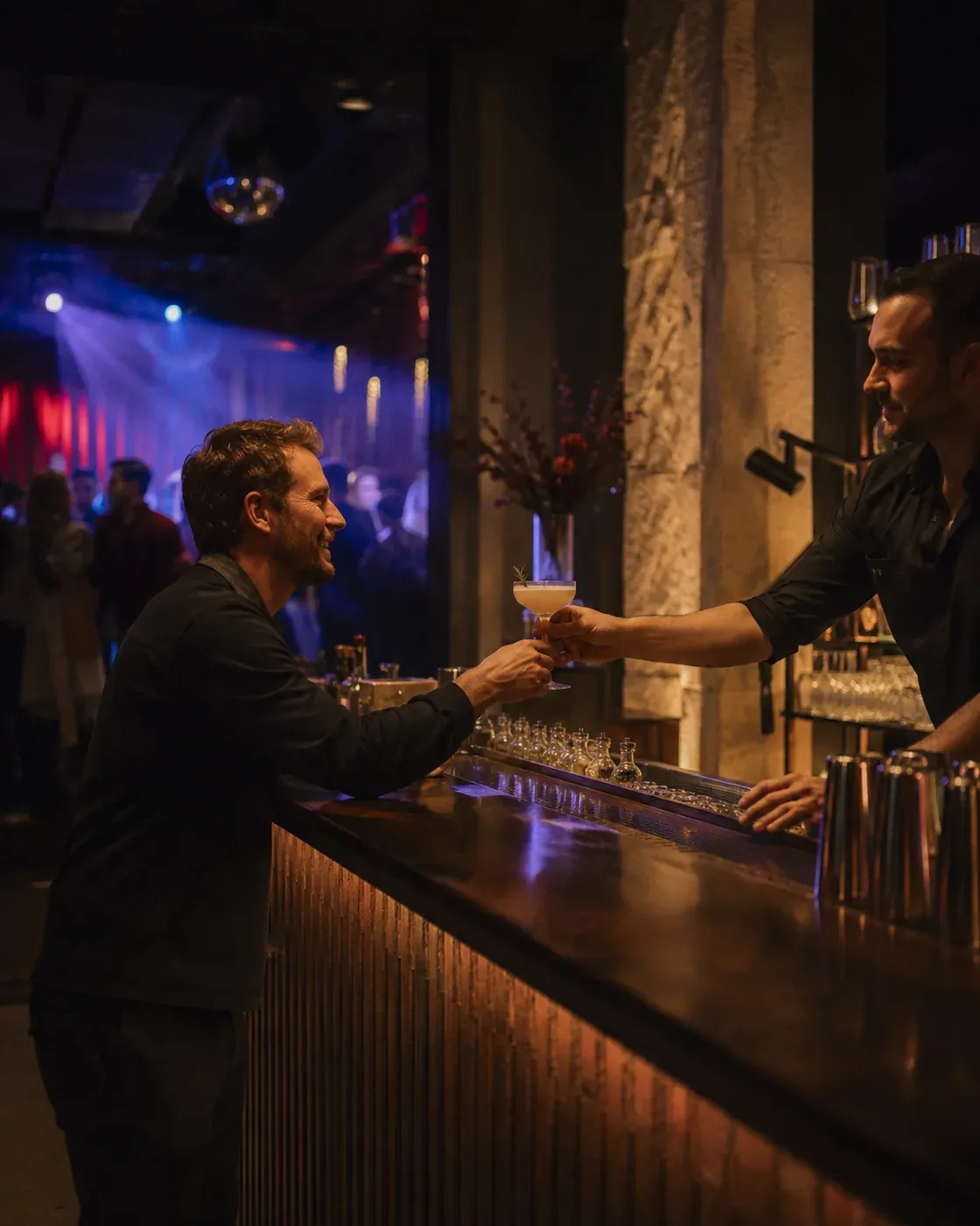 Illustrative scene of a bartender handing a drink to a guest in a nightclub, with the dance floor and stage lighting behind them.