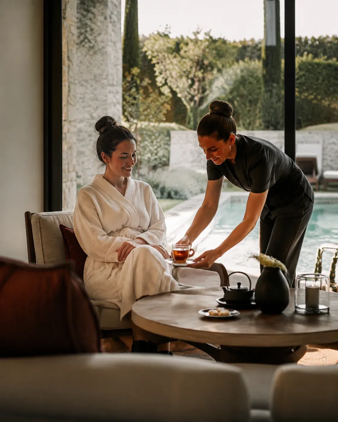 Illustrative scene of a team member bringing tea to a guest in a robe in a quiet spa lounge beside the pool.